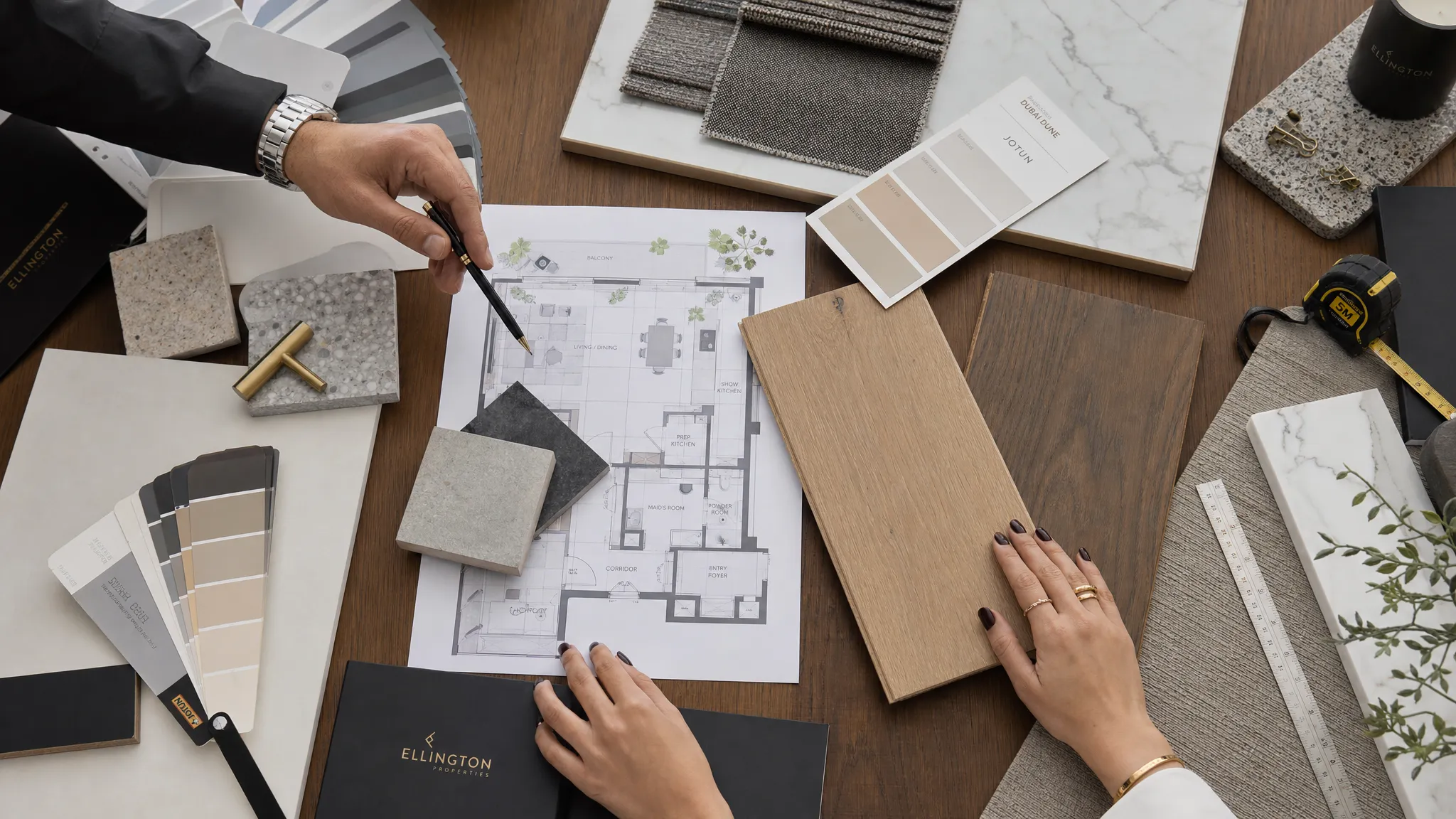 A close-up overhead view of a renovation consultation table with interior material samples, floor plans, wood finishes, paint swatches, tile pieces, and measuring tools arranged neatly for a Dubai home renovation planning meeting.