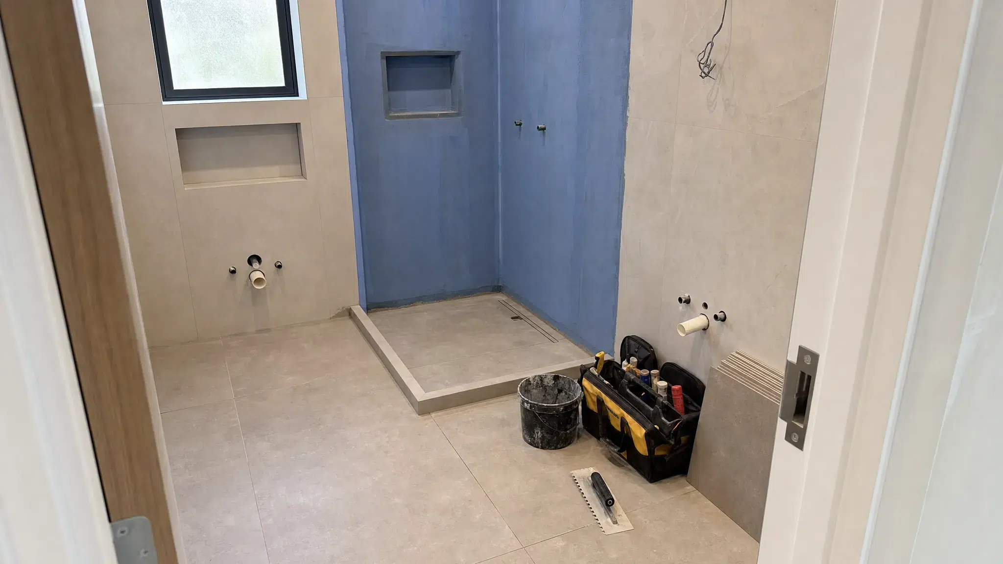 A modern bathroom renovation in progress with new floor tiles, exposed plumbing points, waterproofed shower walls and neatly organized tools, shown from a doorway angle in a compact room.
