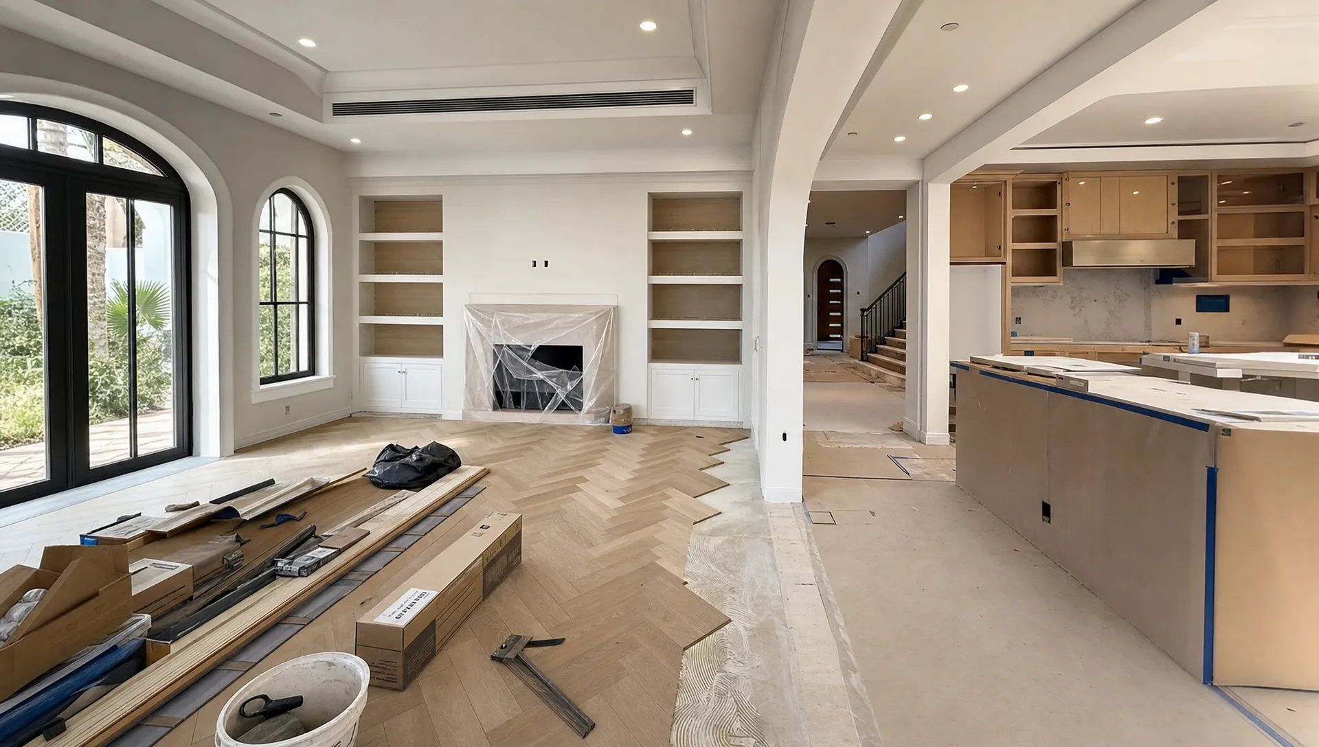 A Dubai villa living area undergoing a coordinated renovation, with new flooring being installed, freshly painted walls, carpentry work in progress, and an open kitchen area prepared for final finishes, seen from a corner angle with materials arranged across the room.