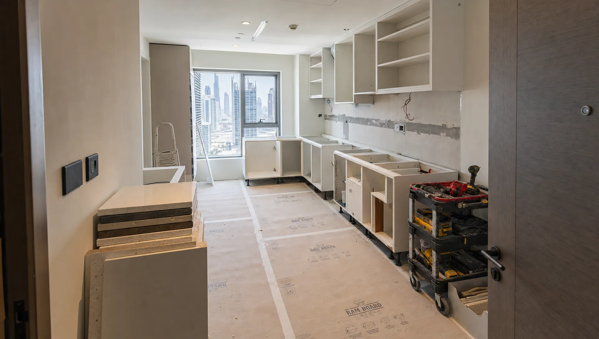 A Dubai apartment kitchen under renovation with cabinet carcasses being fitted, protected flooring, stacked countertop samples, and organized tools on the side, seen from the doorway.