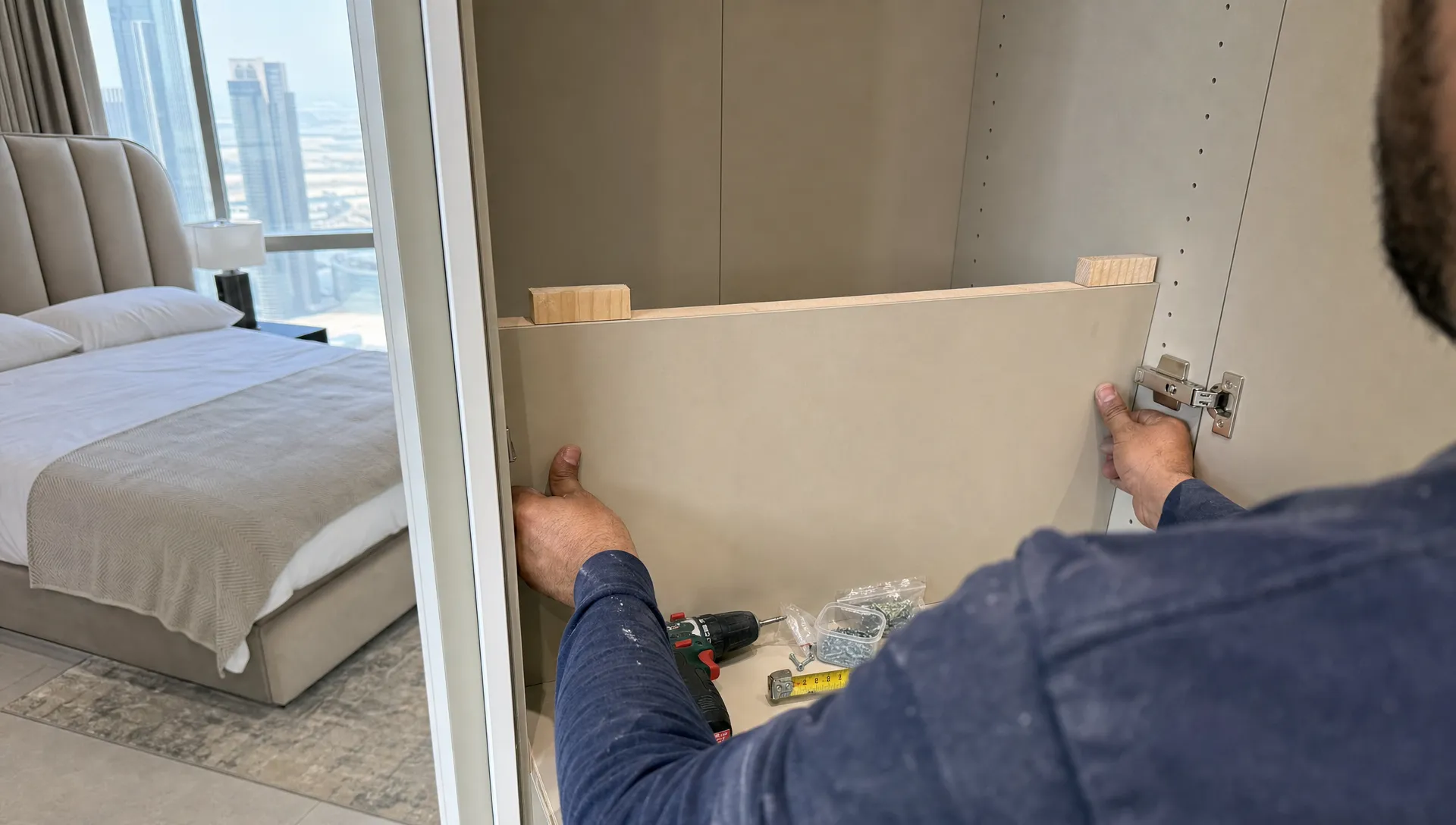 Hands install built-in bedroom storage in a Dubai home, with tools, spacer blocks, and an open wardrobe frame in view.
